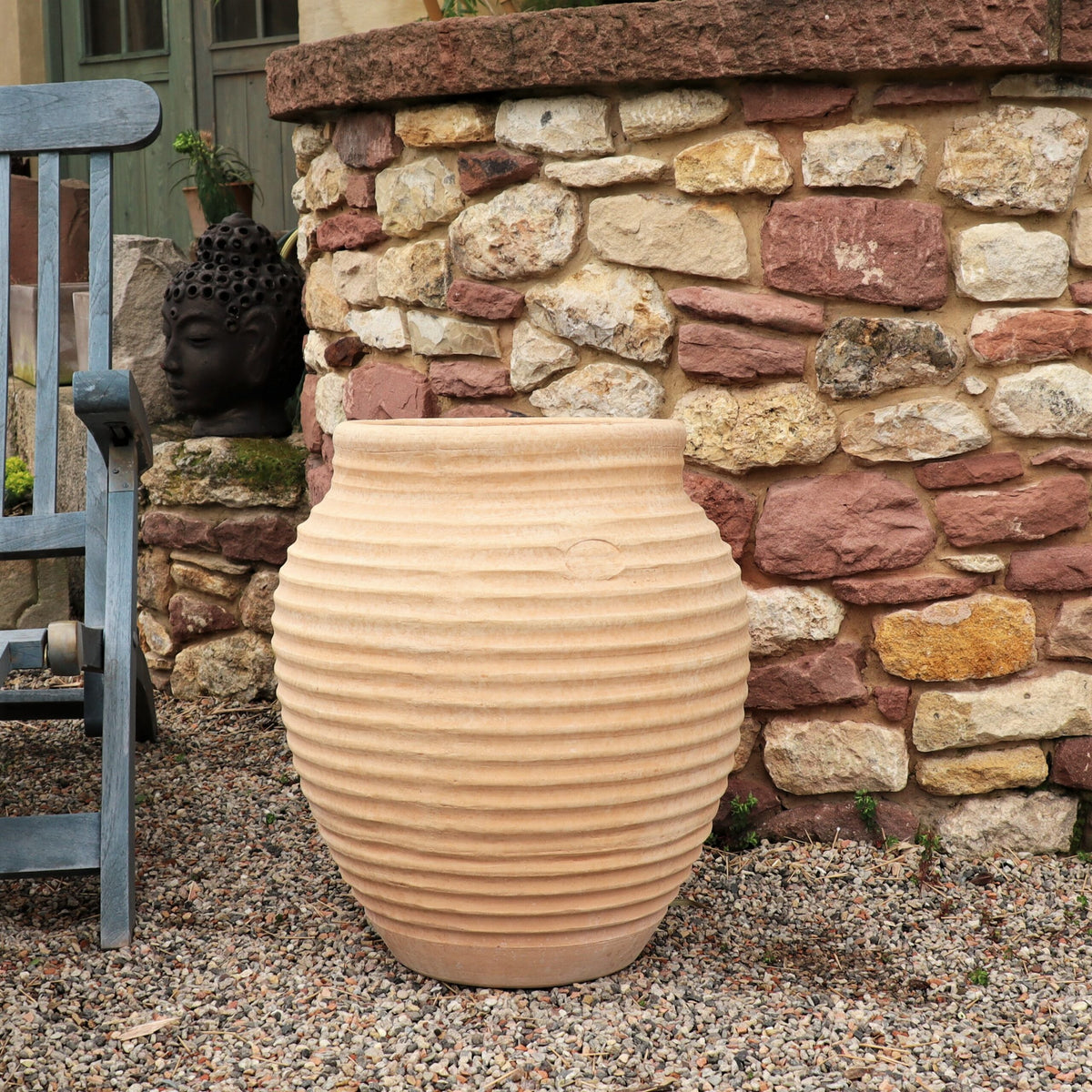 Ein großer, gerippter Terrakotta-Topf steht auf einer Kiesfläche neben einer rustikalen Steinmauer. Daneben stehen ein Holzstuhl und eine dekorative Statue eines ruhigen Buddha-Kopfes, die der Außenumgebung einen Hauch von Ruhe verleihen.