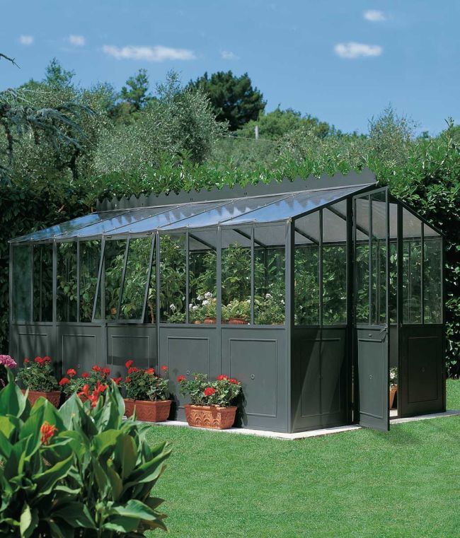 Ein Gewächshaus mit grünem Metallrahmen und großen Glasflächen steht in einem grasbewachsenen Garten. Verschiedene Topfpflanzen und Blumen sind außerhalb des Gewächshauses angeordnet, das von üppigen Bäumen und Büschen unter einem klaren blauen Himmel umgeben ist. Die Gewächshaustür ist geöffnet.