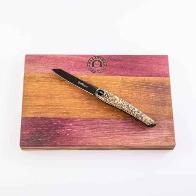 Ein Nesmuk Janus Klappmesser mit verschiedenen Griffmaterialien ruht auf einem handgefertigten Fassholzbrett in drei verschiedenen Farbtönen: Dunkelrot, Hellbraun und Mittelbraun. Das Schneidebrett hat oben in der Mitte ein Emblem mit einem Logo.