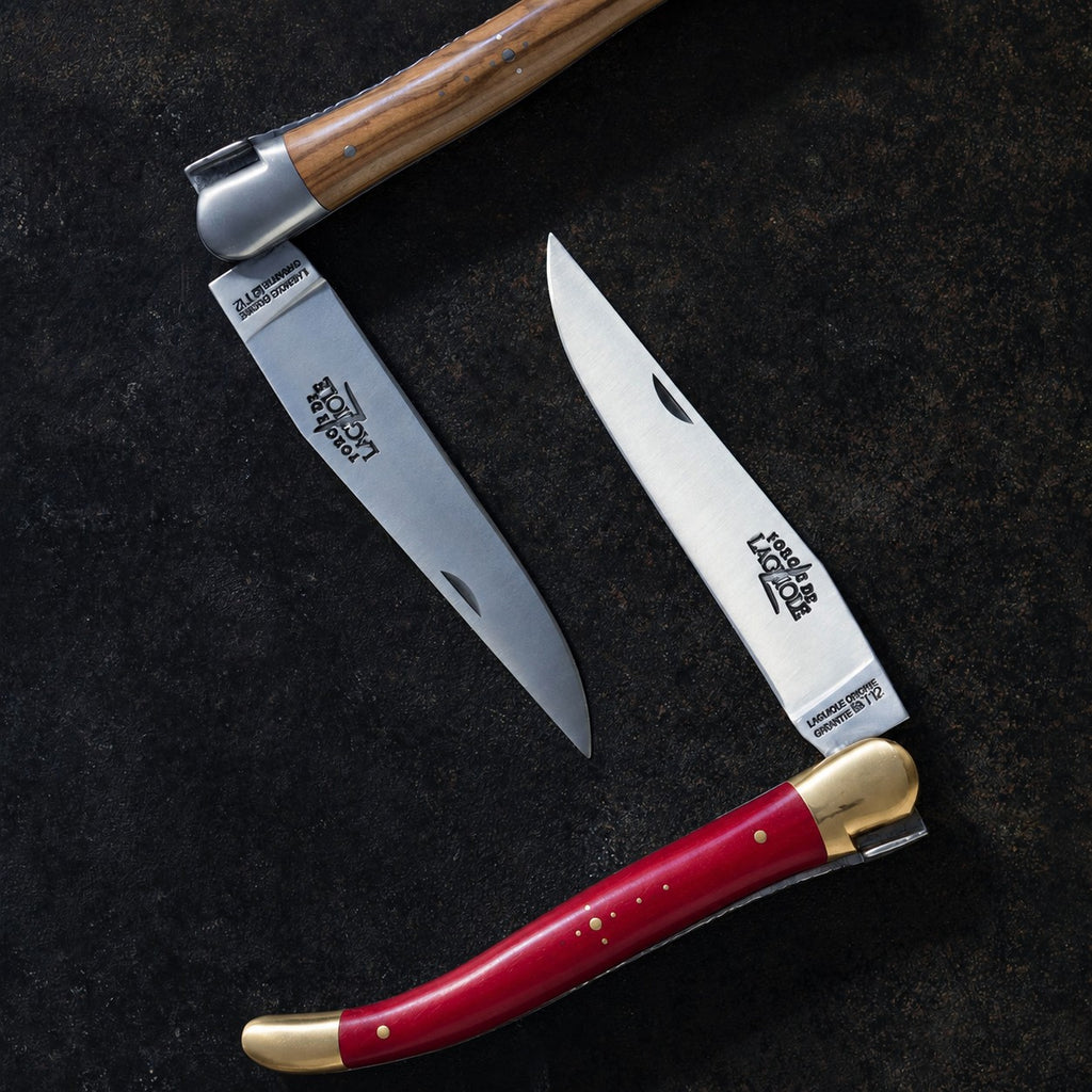 Zwei Klappmesser, eines mit Holzgriff und eines mit rotem Griff, liegen mit ausgeklappten Klingen auf einer dunklen Fläche und bilden dabei die Form des Buchstabens K.
