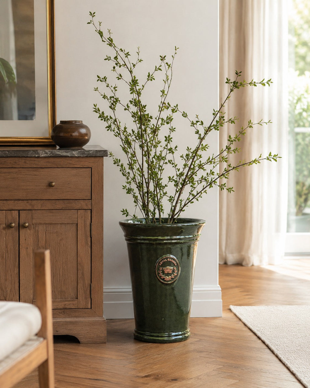 Eine hohe, grüne Vasenform „Garden Green“ (33–51 cm) mit dünnen Zweigen und kleinen Blättern steht auf einem Holzboden neben einem Schrank in einem sonnendurchfluteten, gemütlichen Raum mit transparenten Vorhängen.