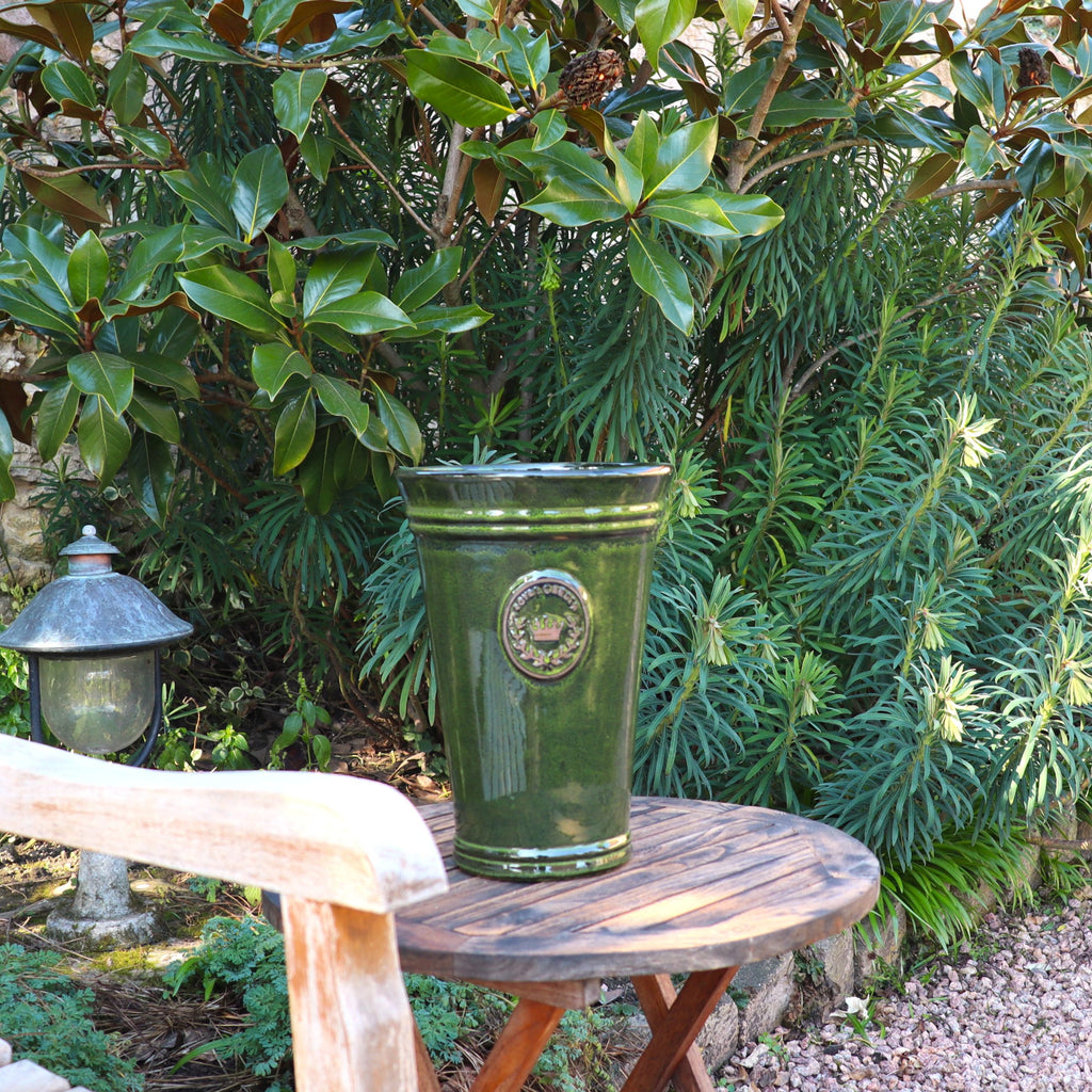 Eine hohe, glänzend grüne Keramikvase aus der Vasenform „Garden Green“ (Höhe 33–51 cm) steht auf einem runden Holztisch in einem Garten, umgeben von üppigen Pflanzen, mit einem Steinweg und einer Außenlampe in der Nähe.