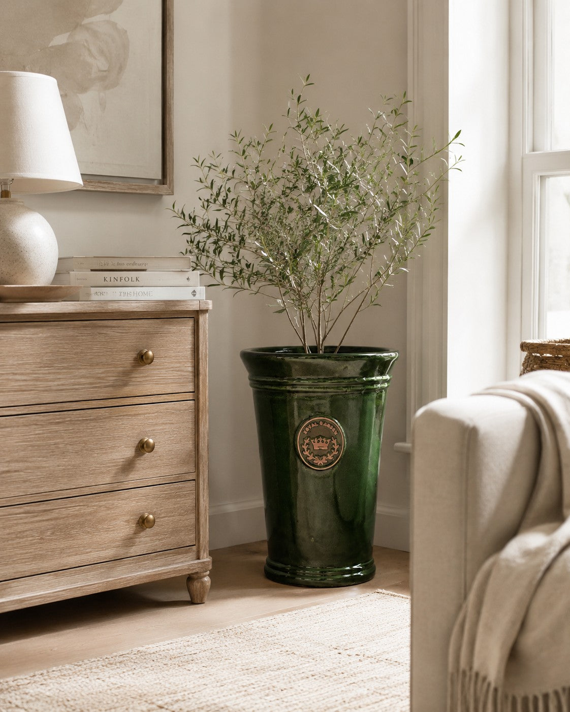 Ein Olivenbaum im Topf, der in einem hohen grünen Keramik-Pflanzgefäß steht – ähnlich dem Modell „Garden Green“ von Vasenform (Höhe 33 bis 51 cm) –, steht neben einer Holzkommode mit Messingknöpfen in einem hellen Wohnzimmer mit neutraler Einrichtung und natürlichem Licht.