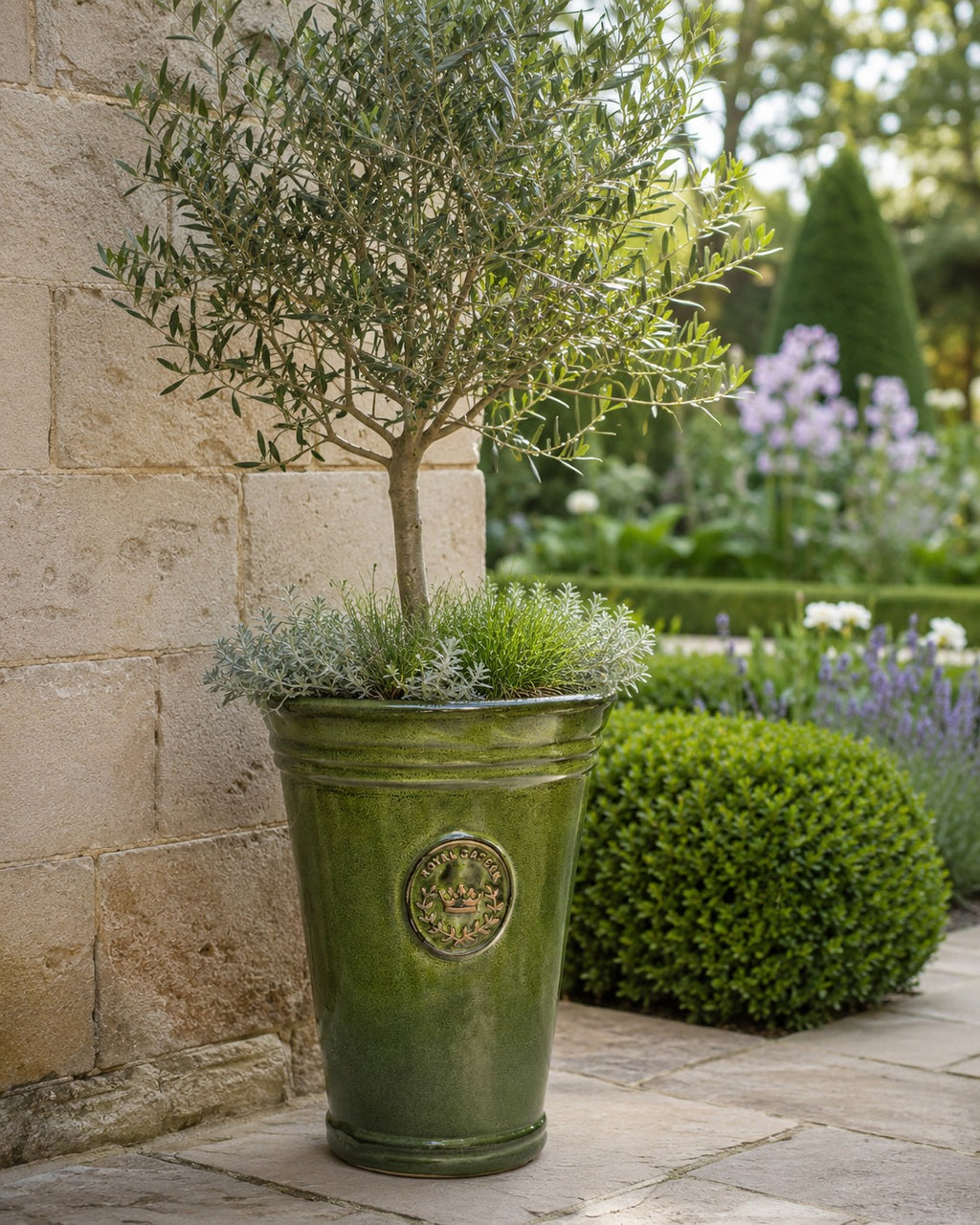 In einem Keramik-Pflanzgefäß der Marke Vasenform in der Farbe „Garden Green“ (Höhe 33 bis 51 cm) stehen ein kleiner Olivenbaum und Blattpflanzen; das Gefäß steht auf einer Steinterrasse neben einer Steinmauer, im Hintergrund ist ein gepflegter Garten zu sehen.