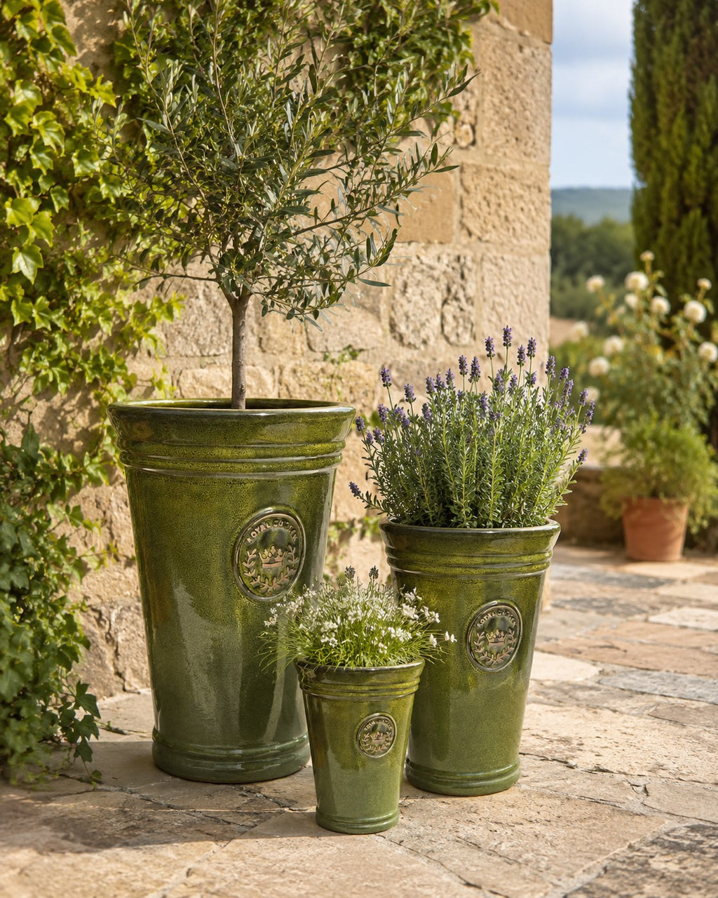 Drei grüne Vasen aus der Vasenform-Serie „Garden Green“ (Höhe 33–51 cm) zeigen einen Olivenbaum, Lavendel und Kräuter auf einer Steinterrasse, arrangiert vor einer rustikalen Steinmauer mit üppigem Grün im Hintergrund.