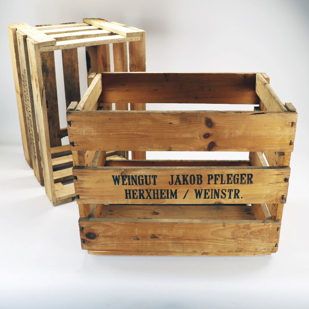 Ausgestellt sind zwei vielseitig einsetzbare Weinkisten im Vintage-Look. Eine Kiste ist auf die Seite gekippt, während die andere aufrecht steht und eine majestätische Präsenz ausstrahlt. Diese Lattenkisten aus verwittertem Holz strotzen vor Charme; eine trägt stolz in schwarzer Schrift die Aufschrift „Weingut Jakob Pfleger Herxheim/Weinstr.“. Jede 48 x 32 x 38 cm große Kiste eignet sich perfekt als Tafelaufsatz und kann als Regal, Schuhregal oder Dekorationselement dienen.