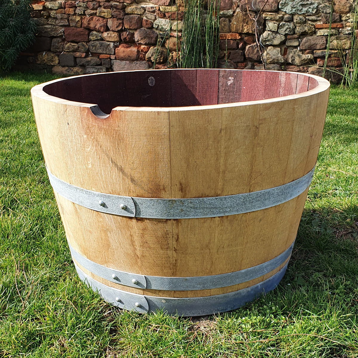 Ein großes halbes Weinfass, Barrique, Holzfass, Blumenkübel mit zwei Metallbändern darum steht aufrecht auf einer Rasenfläche. Das Fass hat oben eine Öffnung und steht vor dem Hintergrund einer rustikalen Steinmauer und hoher, dünner Pflanzen.