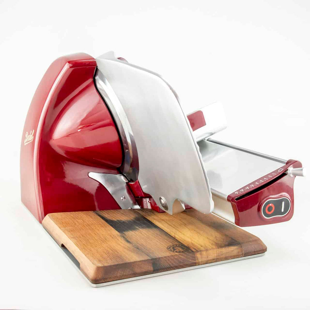 Die Aufschnittmaschine Berkel Home Line 250 Rot mit einem handgefertigten Schneidebrett aus alten Weinfässern ist ein eleganter, roter elektrischer Fleischschneider mit einer glänzenden Metallklinge. Dieser professionelle Allesschneider verfügt über einen Bedienknopf an der Seite und eine einstellbare Schnittstärkenführung, alles vor einem schlichten weißen Hintergrund.