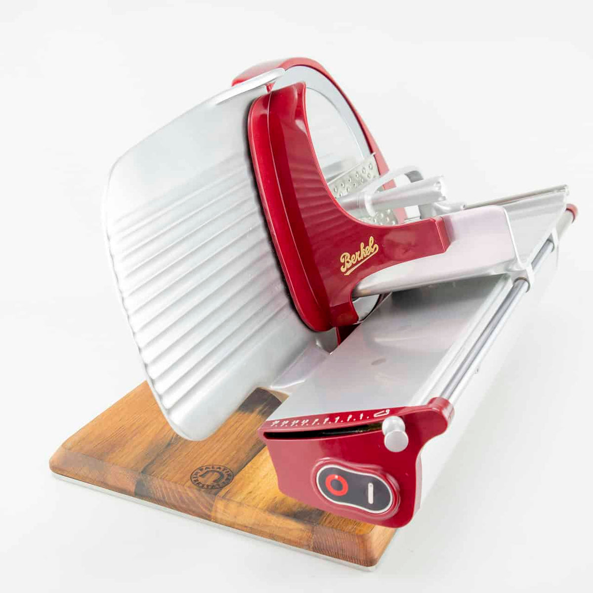 Eine Berkel Home Line 250 Rot Aufschnittmaschine mit einem handgefertigten Holzsockel aus alten Weinfässern. Diese Profi-Aufschnittmaschine verfügt über eine scharfe, gezahnte Klinge, die durch einen Metallschutz und einen einstellbaren Dickenknopf geschützt ist. Der Markenname „Berkel“ ist auf dem rot-silbernen Gehäuse deutlich sichtbar. Der Hintergrund ist schlicht weiß.