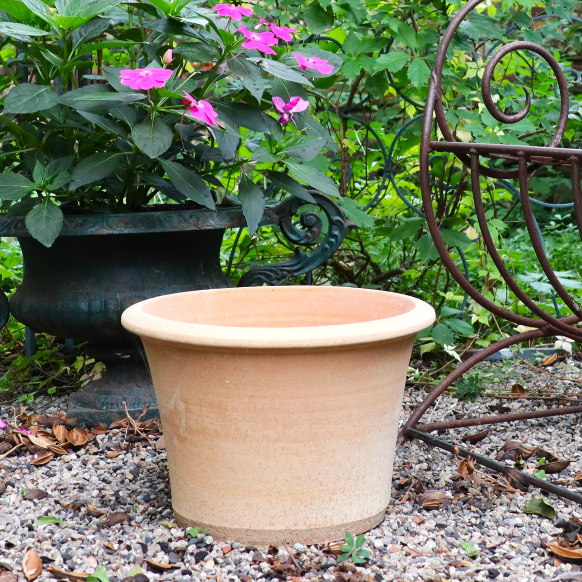 Der handgemachte Terrakotta Pflanztopf "Hosta" 35 cm - frostfest - steht auf Kies in einem Garten neben einem Metallstuhl und einem dunklen Übertopf mit rosa Blüten, umgeben von üppigem Grün.