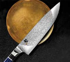 Eine Nahaufnahme des limitierten Kochmessers KAI SHUN Engetsu TA-0706, eines von nur 842 erhältlichen Exemplaren, mit einer markanten 20 cm langen Damastklinge vor einem goldbraunen, kreisförmigen Hintergrund auf einer schwarzen Oberfläche. Das Messer hat einen dunklen Griff und die Klinge weist ein kompliziertes Muster auf.