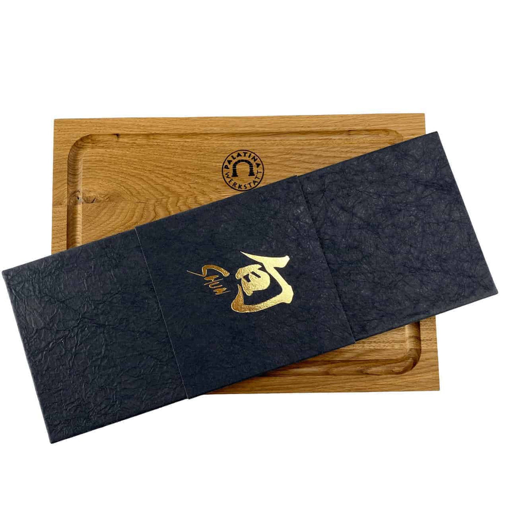 Ein 30x22 cm großes Schneidebrett aus Eiche mit vertiefter Mitte. Darauf liegt ein geschlossenes, strukturiertes schwarzes Etui mit goldenen japanischen Schriftzeichen und einem Logo mit einer Person in konzentrischen Kreisen oben links, perfekt zur Präsentation Ihres Kai Shun Classic Chinakochmesser DM-0712.
