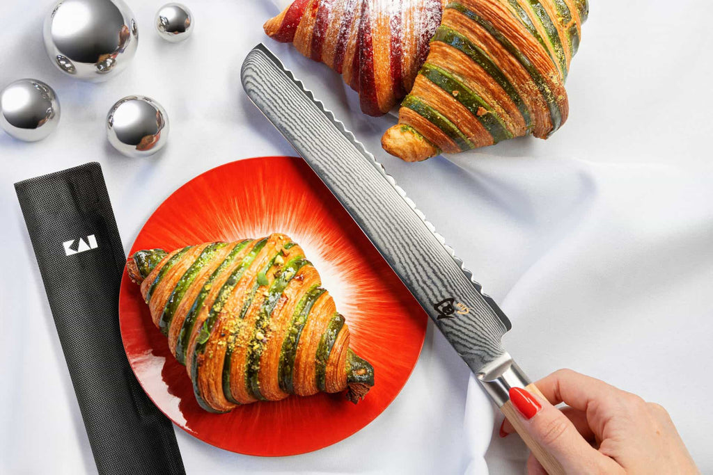 Eine Person hält ein KAI X-MAS SELECTION DREAMBOX Shun Classic White Brotmesser mit Klingenschutz in der Hand, neben zwei bunten Croissants auf einem weißen Tuch. Ein Croissant liegt auf einem roten Teller, das andere ruht daneben, begleitet vom Klingenschutz und drei glänzenden silbernen Kugeln, die die Eleganz der Szene vervollständigen.