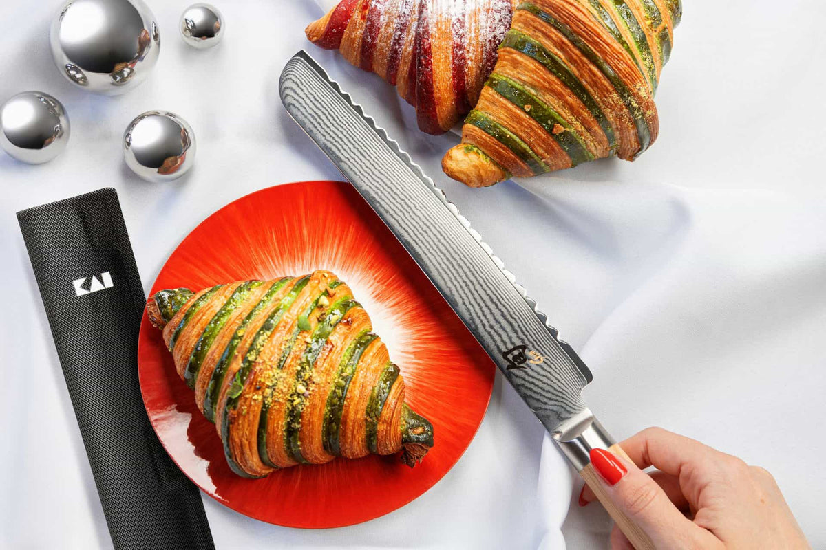 Eine Person hält ein KAI X-MAS SELECTION DREAMBOX Shun Classic White Brotmesser mit Klingenschutz in der Hand, neben zwei bunten Croissants auf einem weißen Tuch. Ein Croissant liegt auf einem roten Teller, das andere ruht daneben, begleitet vom Klingenschutz und drei glänzenden silbernen Kugeln, die die Eleganz der Szene vervollständigen.