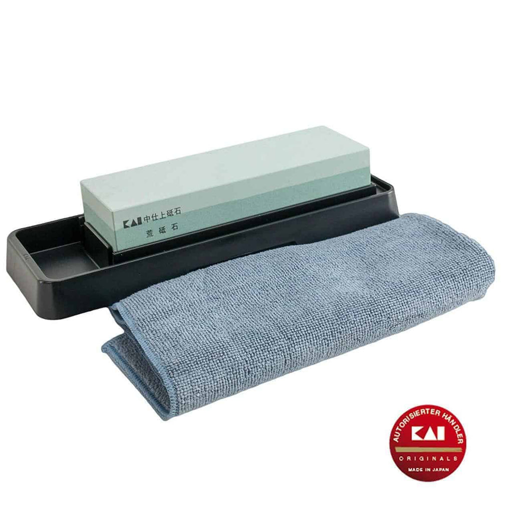 Ein rechteckiger Kai AP-0305 Schleifstein | Wetzstein Messerschärfer 400/1000 Körnung mit schwarzer Basis, daneben liegt ein gefaltetes blaues Poliertuch. Der Schleifstein trägt japanische Inschriften und in der unteren rechten Ecke befindet sich ein roter runder Aufkleber mit der Aufschrift „KAI“ und „Made in Japan“.