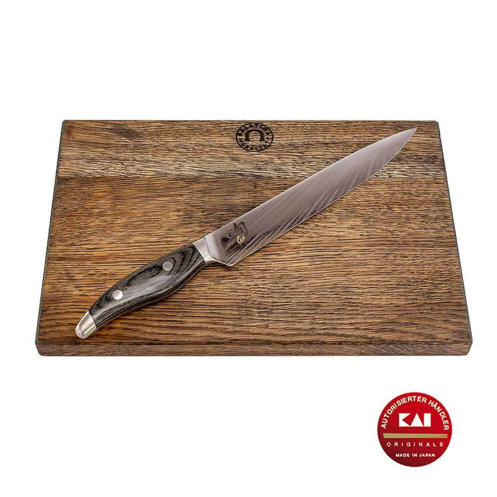 Ein schlankes Kai Shun Nagare NDC-0704 ultrascharfes Schinkenmesser mit dunklem Holzgriff liegt auf einem basaltgrauen Schneidebrett 34x21 cm (Eiche). Die 23 cm lange Klinge aus 72 Lagen Damaststahl weist ein markantes Muster auf, und das Schneidebrett trägt oben in der Mitte ein Logo. Ein rotes Siegel in der unteren rechten Ecke weist darauf hin, dass es in Japan von Kai hergestellt wurde.