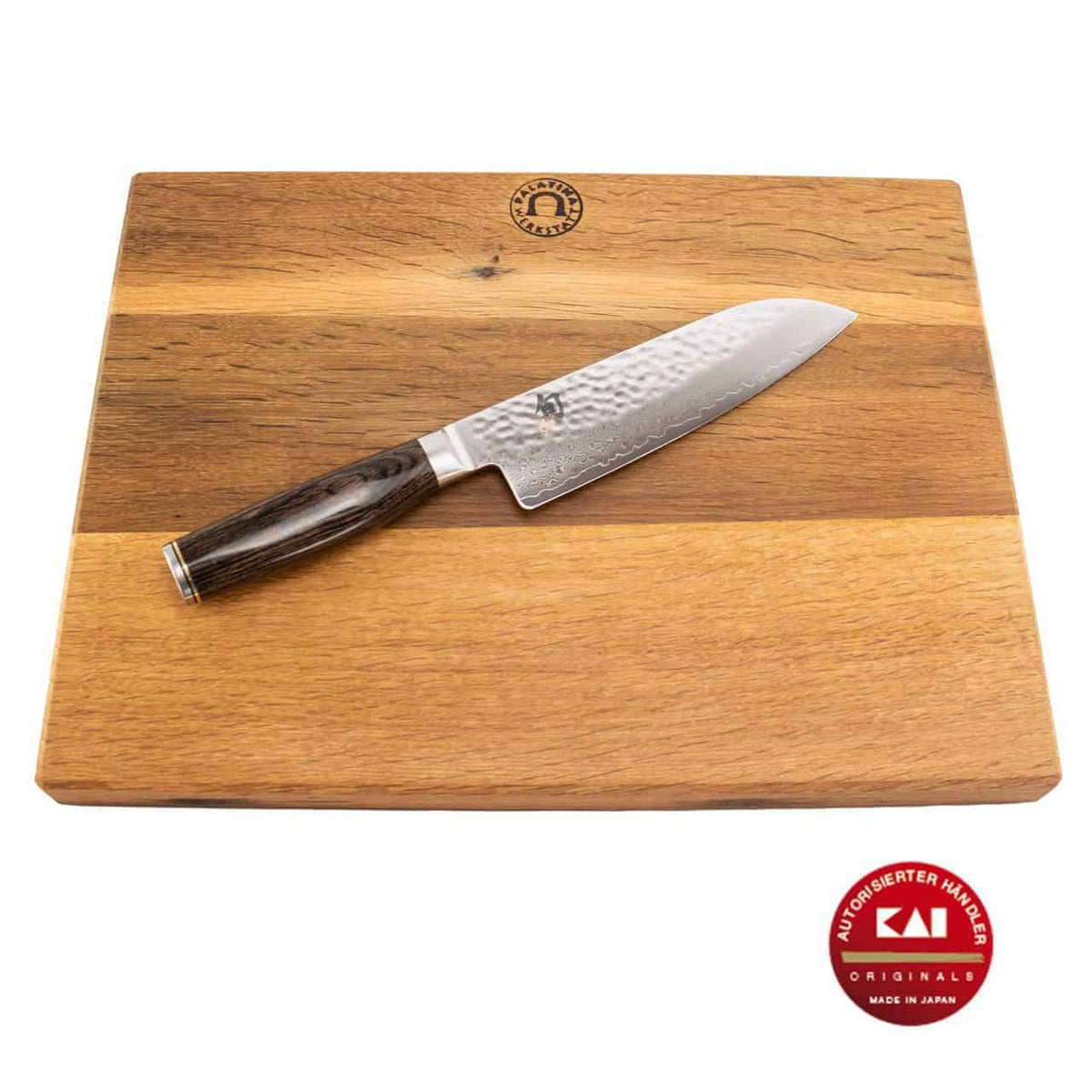 Ein Kai Shun Tim Mälzer TDM-1702 Santokumesser mit 18 cm langer Damaszenerstahlklinge und dunklem Griff ruht diagonal auf einem 40x30 cm großen Küchenarbeitsbrett aus Holz. Die Logos des Schneidebrettherstellers und des Messerherstellers sind deutlich zu erkennen. Das Santokumesser trägt die Aufschrift „Kai Original“ und „Made in Japan“.