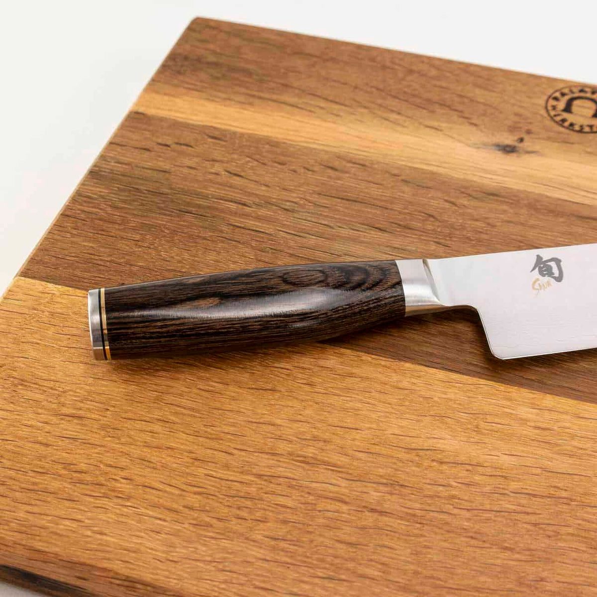 Ein hochwertiges Kai Shun Tim Mälzer TDM-1702 Santokumesser mit dunklem Holzgriff ruht auf einem hölzernen Küchenarbeitsbrett 40x30 cm. Die 18 cm lange Damaststahlklinge hat eine reflektierende Oberfläche und ist mit einem kleinen aufgedruckten Emblem in der Nähe des Griffs versehen. Das Schneidebrett weist eine natürliche Holzmaserung mit unterschiedlichen Schattierungen auf.