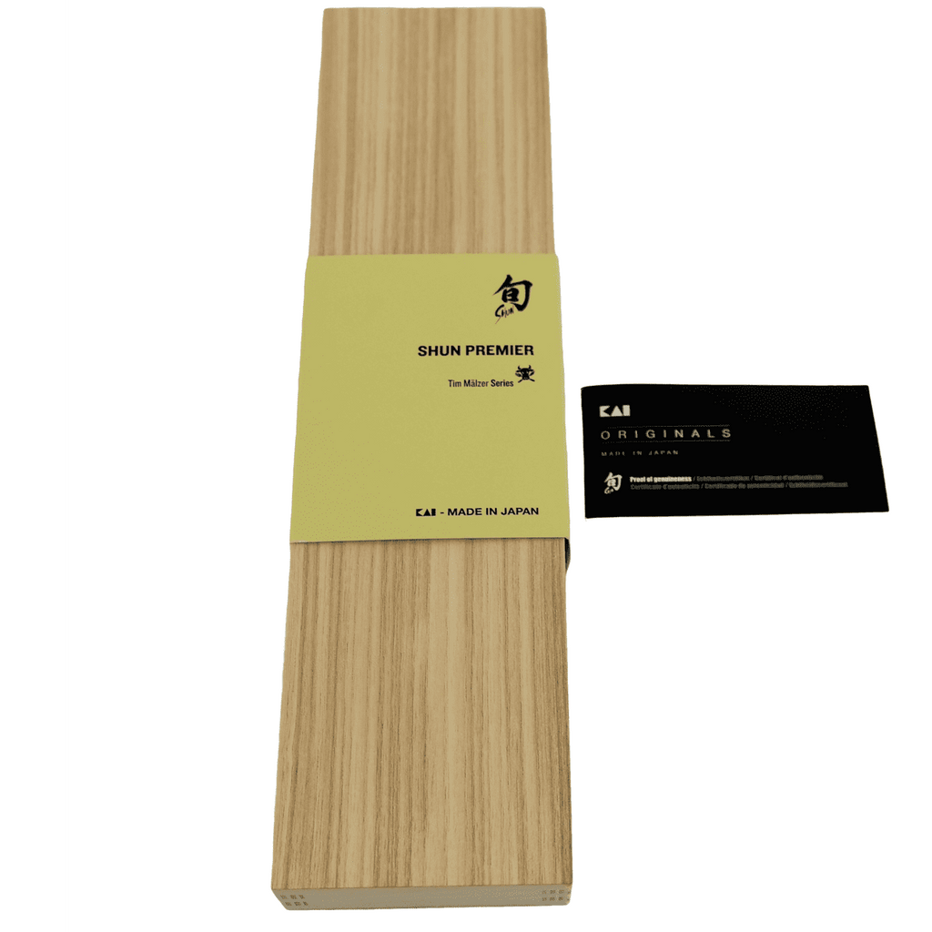 Eine rechteckige Holzbox mit einem hellgelben Band mit der Aufschrift „Shun Premier Tim Mälzer Series“ in schwarzer Schrift. Das Kai Shun Premier Geschenkset wird von einer schwarzen Karte mit weißer Schrift und einem Logo begleitet, das darauf hinweist, dass es von KAI und in Japan hergestellt wird. Es enthält exquisites Besteck wie das TDM-1715 Tourniermesser mit einer 5,5 cm langen Klinge und wird mit einem messerfreundlichen Schneidebrett aus Fassholz (Eiche) mit den Maßen 25 x 15 cm geliefert.