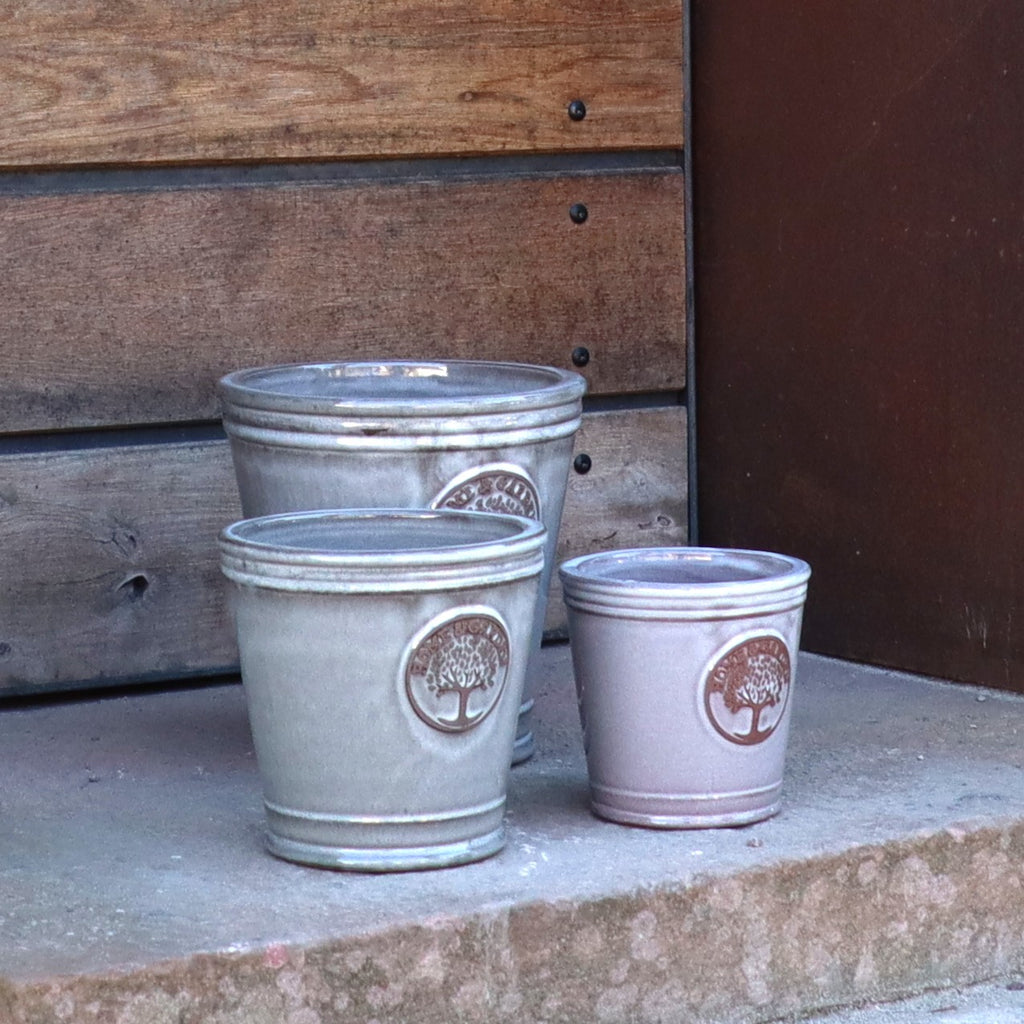 Drei leere Keramiktöpfe unterschiedlicher Größe, jeder mit einem Baum-Emblem, stehen auf einer Betonstufe vor Holz und dunklem Metall. Einer davon ist der kleinere, glasierte Pflanzkübel „Cappuccino“ (Höhe 15–24 cm), der dem Gesamtbild einen dezenten Farbtupfer verleiht.