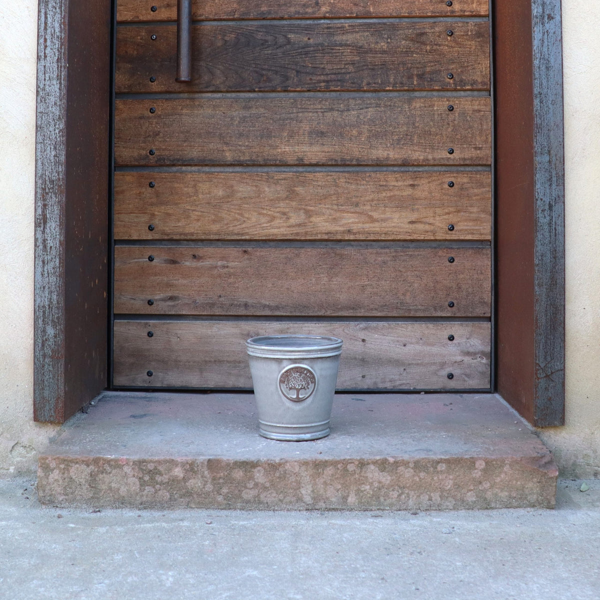 Ein kleiner Metalleimer steht auf einer Betonschwelle vor einer großen Holztür mit horizontalen Brettern und einem Metallgriff, neben dem kleinen, glasierten Pflanzkübel „Cappuccino“ (Höhe 15–24 cm).