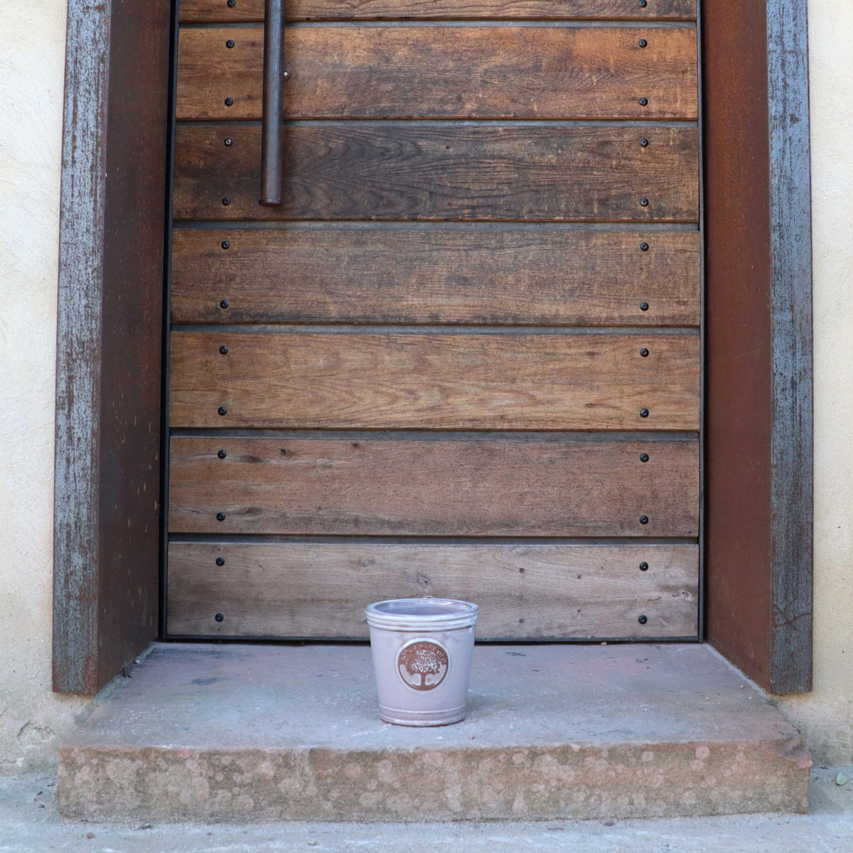 Ein kleiner, glasierter Pflanzkübel „Cappuccino“ (Höhe 15–24 cm) mit Logo steht auf einer Betontreppe vor einer großen, verwitterten Holztür mit Eisenbeschlägen.