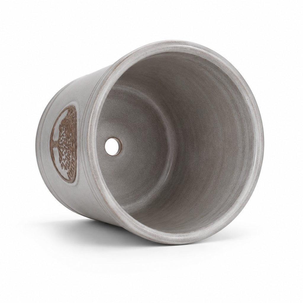 Der kleine, glasierte Pflanzkübel „Cappuccino“ (Höhe 15–24 cm) liegt auf der Seite, sodass man das Innere und ein Abflussloch am Boden sehen kann; seine graue Außenseite ist mit einem dezenten metallischen Ziermuster verziert.