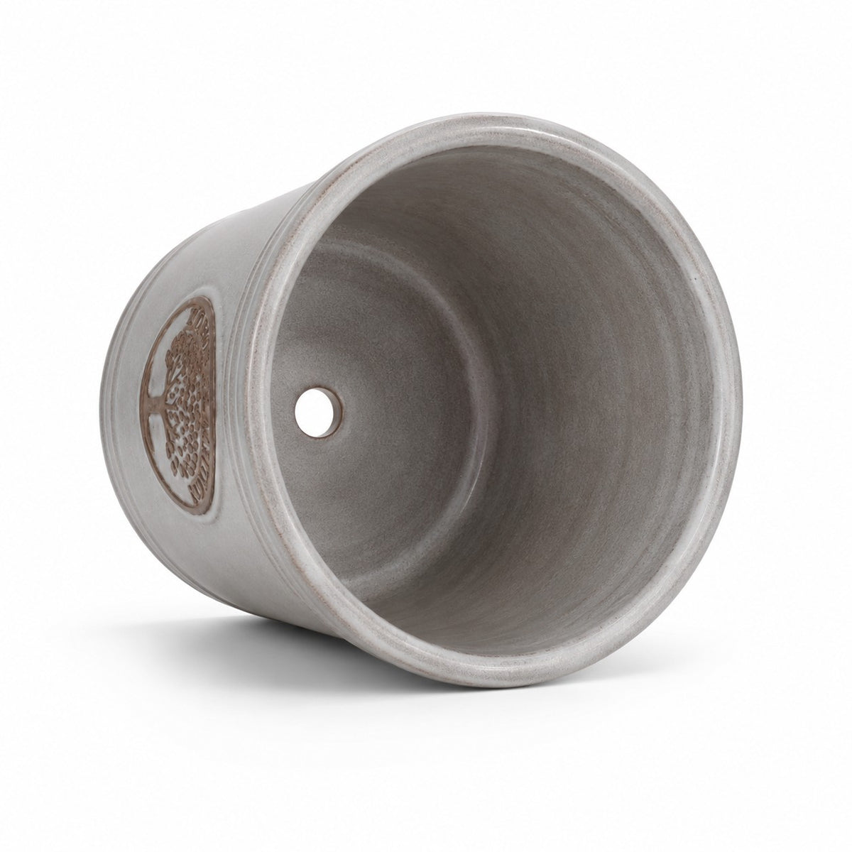 Der kleine, glasierte Pflanzkübel „Cappuccino“ (Höhe 15–24 cm) liegt auf der Seite, sodass man das Innere und ein Abflussloch am Boden sehen kann; seine graue Außenseite ist mit einem dezenten metallischen Ziermuster verziert.