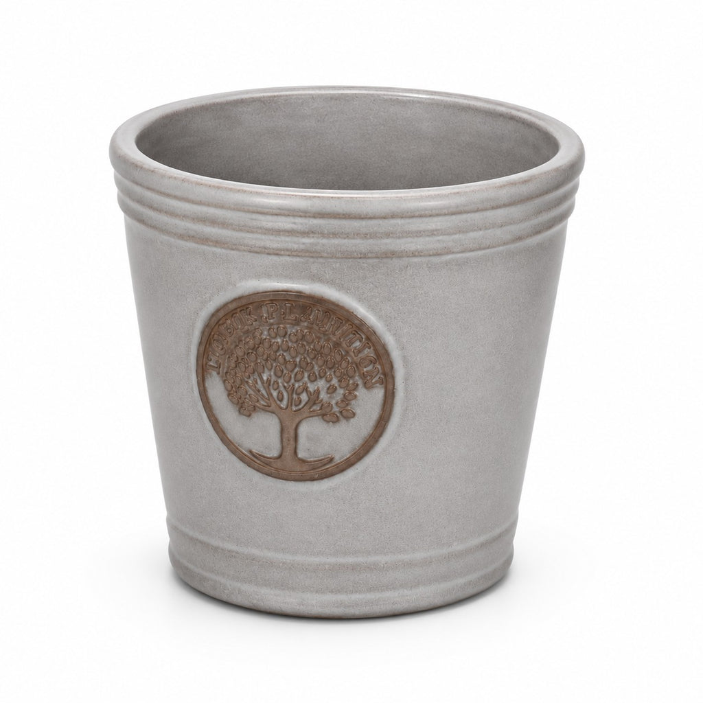 Der kleine, glasierte Pflanzkübel „Cappuccino“ (Höhe 15–24 cm) in Grau mit Baum-Emblem und dezenten Rillen am Rand und am Sockel verleiht mit seinem metallischen Schimmer vor weißem Hintergrund ein elegantes Flair.