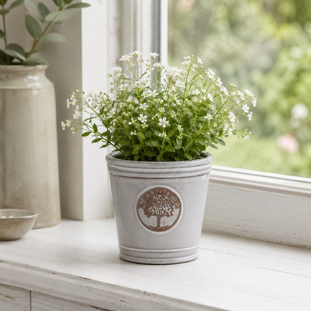 Ein kleiner, glasierter Pflanzkübel „Cappuccino“ (Höhe 15–24 cm) mit Baum-Emblem steht auf einer weißen Fensterbank und ist mit zarten weißen Blüten bepflanzt. Draußen ist Grün zu sehen, im Hintergrund ein Teil einer Keramikvase.