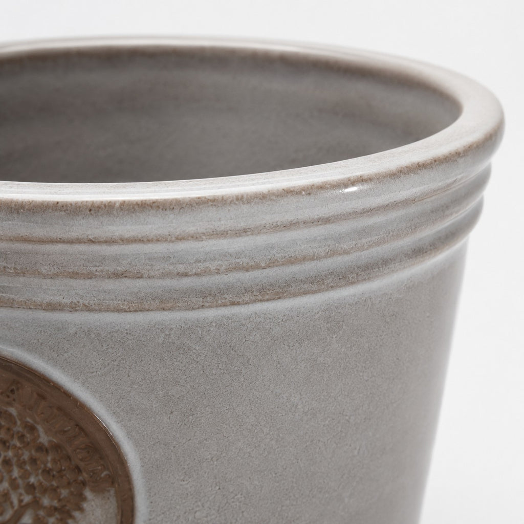 Nahaufnahme des oberen Randes und der Seite des kleinen, glasierten Pflanzkübels „Cappuccino“ (Höhe 15–24 cm) in Hellgrau mit leicht glänzender Oberfläche und einem erhabenen, kreisförmigen Emblem an der Seite, das einen dezenten metallischen Schimmer verleiht.