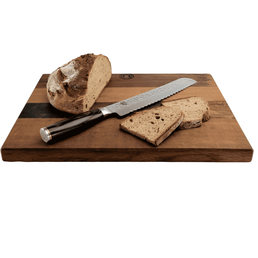 Ein Laib rustikales Brot und mehrere Scheiben sowie ein KAI Shun Premier Tim Mälzer Geschenkset | TDM-1705 Brotmesser mit dunklem Griff liegen auf einem Holzschneidebrett. Das robuste Fassholzbrett 30x18 cm zeigt ein zweifarbiges Muster mit abwechselnd dunklen und hellen Holzstreifen.