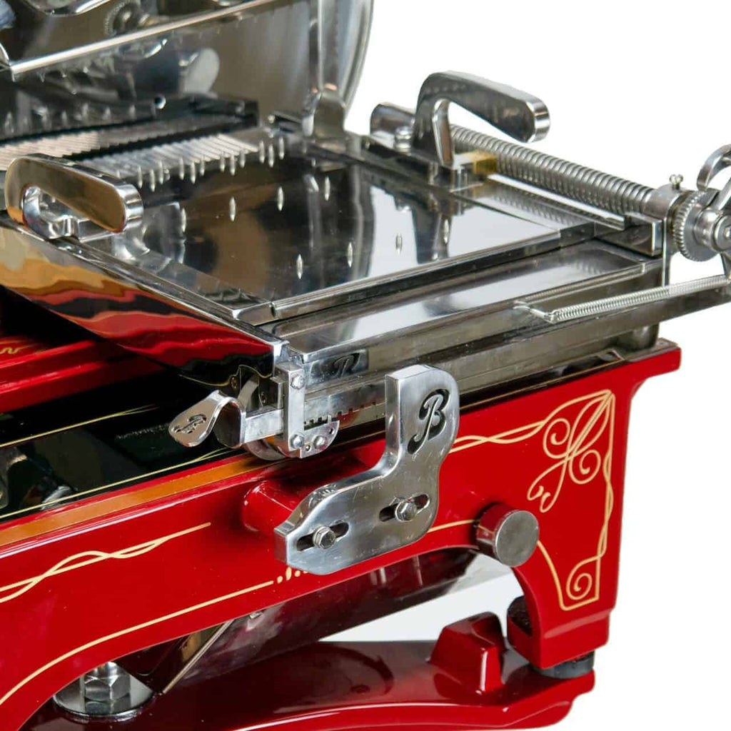Nahaufnahme der roten Berkel Volano L16 nostalgischen Schwungrad-Aufschnittmaschine mit silbernen Akzenten und handgefertigtem Einlegebrett. Der Allesschneider ist mit kunstvollen Details versehen, darunter dekorative Schnörkel. Die Klinge und der Schneidemechanismus sind sichtbar und zeigen das komplexe Design und die Handwerkskunst.
