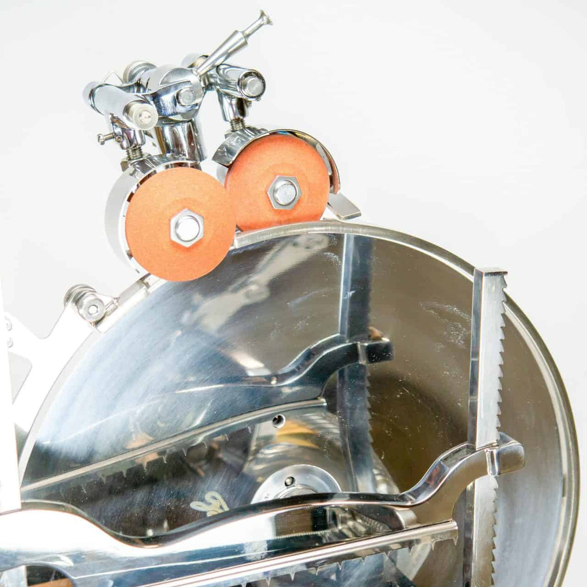 Eine Nahaufnahme einer glänzenden, verchromten Berkel Volano L16 nostalgischen Schwungrad-Aufschnittmaschine rot + handgefertigtem Einlegebrett mit sichtbaren Klingen und angebrachten orangefarbenen Schleifsteinen. Der Aufschnitter hat eine reflektierende Oberfläche und verschiedene mechanische Teile, die eine industrielle und präzise Ästhetik erzeugen.