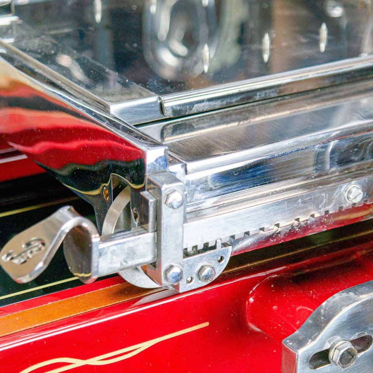Nahaufnahme einer nostalgischen Schwungrad-Aufschnittmaschine Berkel Volano L16 in Rot und Chrom mit komplizierten beweglichen Teilen und strukturiertem Griff. Die Maschine weist glänzende Metallkomponenten und eine reflektierende Oberfläche auf, was darauf schließen lässt, dass es sich um einen Teil eines größeren Geräts oder einer antiken Maschine handeln könnte.