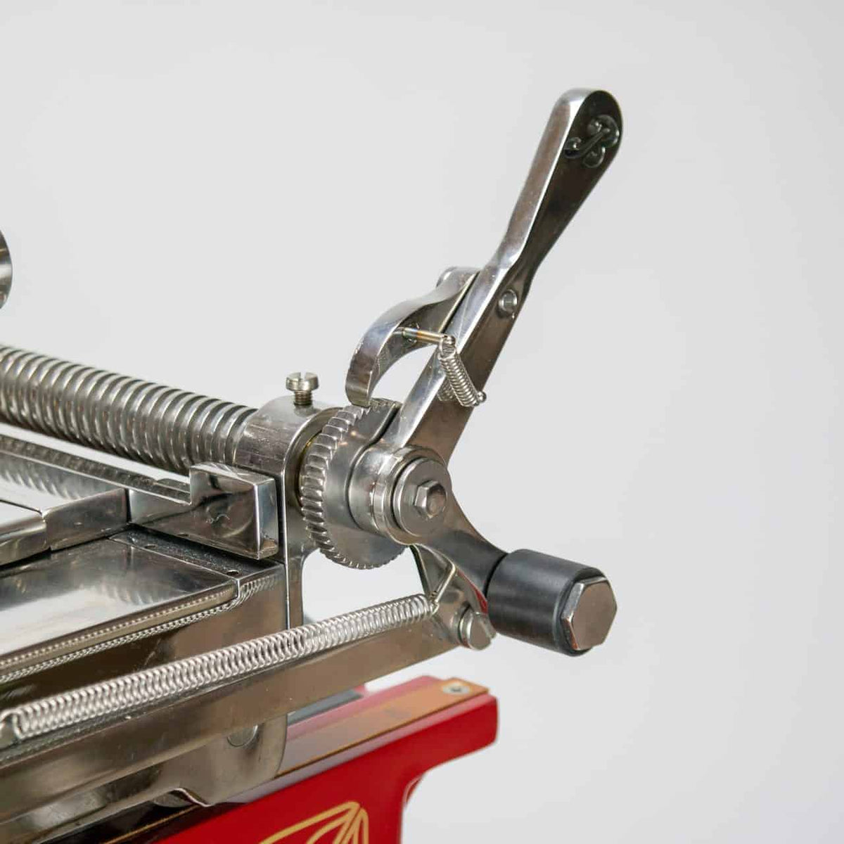 Nahaufnahme eines alten mechanischen Geräts mit Hebel, Zahnrädern und Federmechanismus. Das Gerät ähnelt mit seinem nostalgischen Design der Berkel Volano L16 nostalgische Schwungrad-Aufschnittmaschine rot + handgefertigtem Einlegebrett, besteht aus Metall mit komplizierten Teilen und ist auf einem roten Sockel mit dekorativen Details montiert. Der Hintergrund ist schlicht und weiß.
