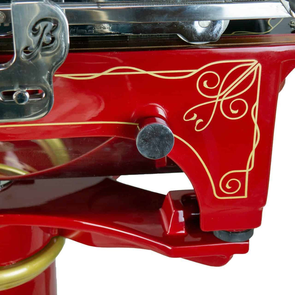Nahaufnahme einer roten Berkel Volano L16 nostalgische Schwungrad-Aufschnittmaschine mit aufwendigen Golddetails am Gehäuse. Der verchromte Einstellhebel und -knopf sind sichtbar und bilden einen auffälligen Kontrast zum Rot. Die Maschine strahlt mit ihrem eleganten, kunstvollen Design und den glänzenden Metallakzenten, ergänzt durch ein handgefertigtes Intarsienbrett, ein klassisches, nostalgisches Gefühl aus.