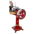 Eine klassische Berkel Volano L16 Schwungradmaschine in Rot mit Chromakzenten auf einem Standfuß. Der Allesschneider verfügt über ein großes Handkurbelrad mit dekorativem Text, eine hölzerne Schneidefläche (Einlagebrett Fassholz) und verschiedene scharfe Metallteile. Der Sockel weist kunstvolle Details auf und steht auf einem vierbeinigen Sockel.
