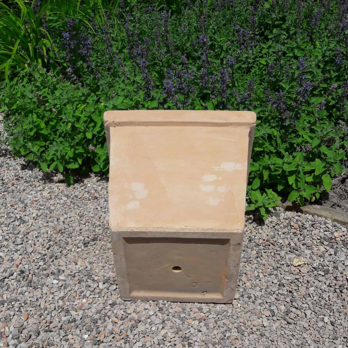 Der Pflanztopf aus Terrakotta „Laurus“, eckig mit einer Höhe von 26 cm, mit einer hellbraunen Terrakotta-Tonstruktur mit schräger Oberseite und einem Loch an der Vorderseite, steht auf einem Kiesweg. Grünes Laub mit violetten Blüten bildet den Hintergrund.