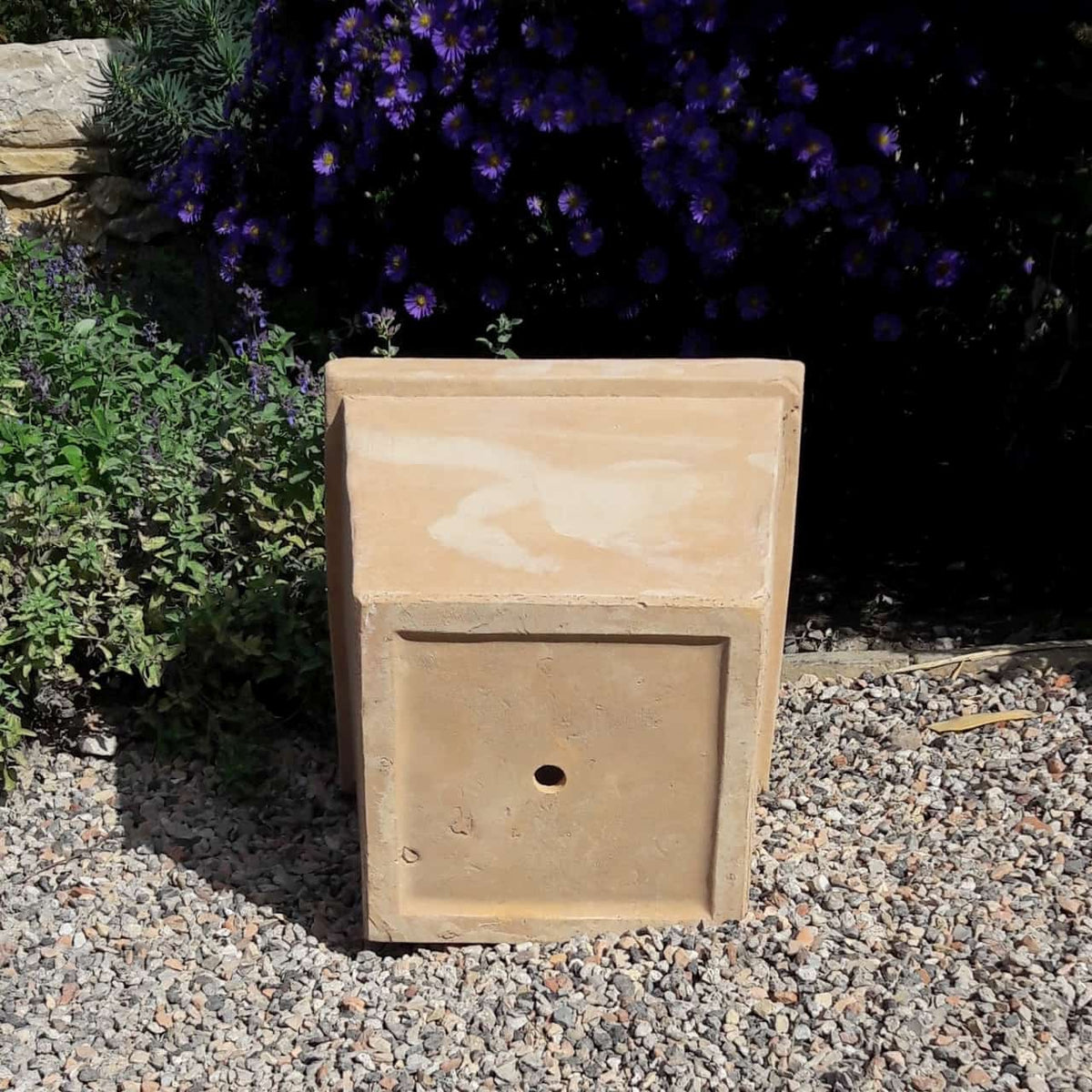 Ein Pflanztopf aus Terrakotta „Laurus“, eckig, H=26cm steht auf einer Kiesfläche, umgeben von grünem Blattwerk, darunter Lorbeersträucher und violette Blumen im Hintergrund. Der Terrakotta-Pflanztopf scheint verwittert und wurde möglicherweise zu dekorativen oder praktischen Zwecken verwendet.
