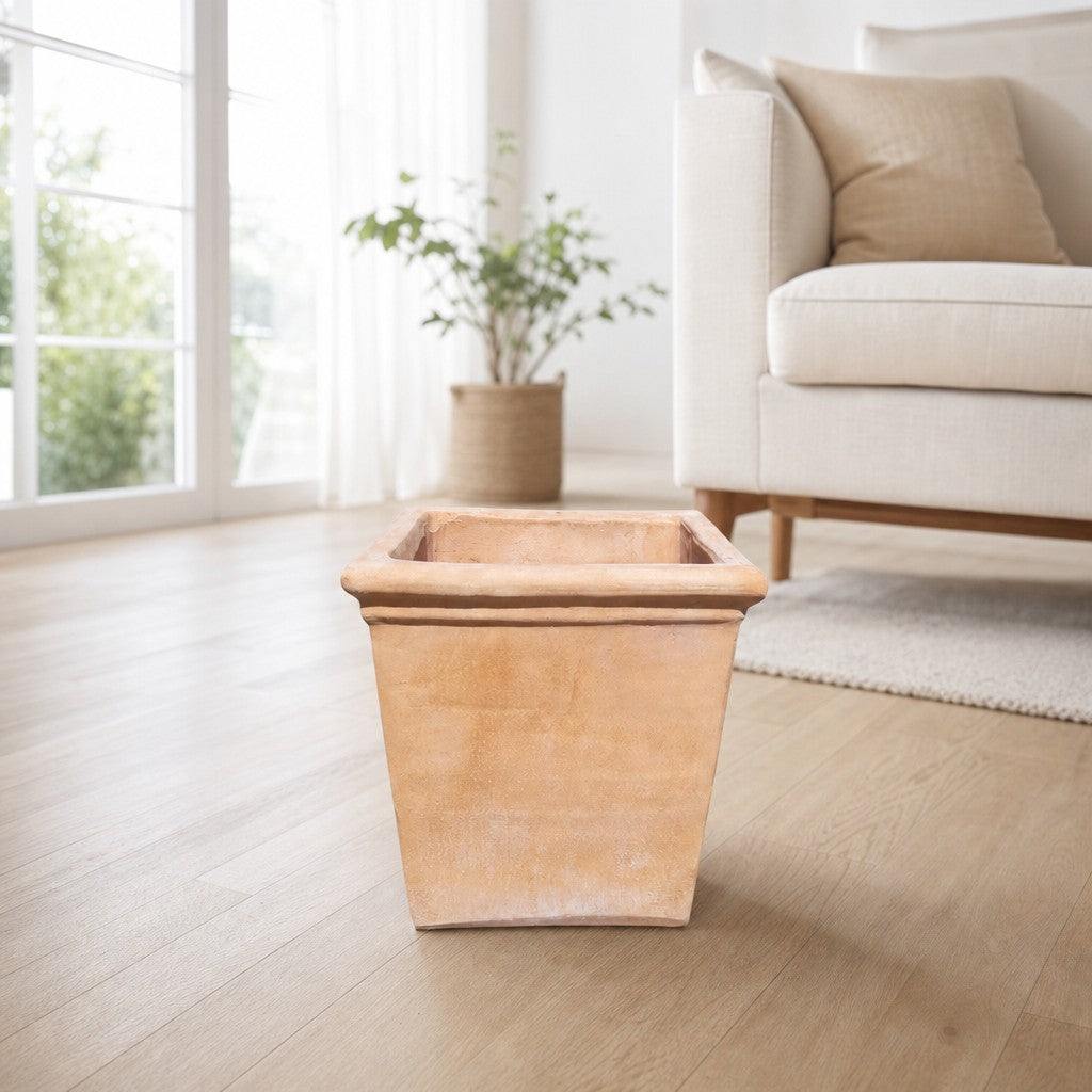 Ein quadratischer Terrakotta-Blumenkübel steht auf einem hellen Holzboden in einem hellen, modernen Wohnzimmer mit einem beigen Sofa, einem strukturierten Teppich und einer Topfpflanze in der Nähe großer Fenster.