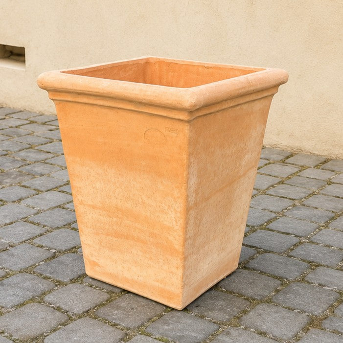Ein großer, leerer, quadratischer Terrakotta-Pflanzkübel steht auf einer mit Steinen gepflasterten Fläche vor einer beigen Wand.