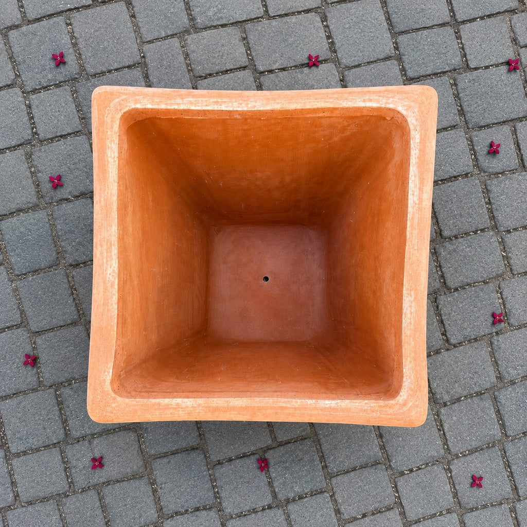 Eine Ansicht von oben auf einen leeren, quadratischen Terrakotta-Blumenkübel mit einem Abflussloch am Boden, der auf grauen, quadratischen Steinfliesen steht, um die herum kleine rosa Blüten verstreut sind.