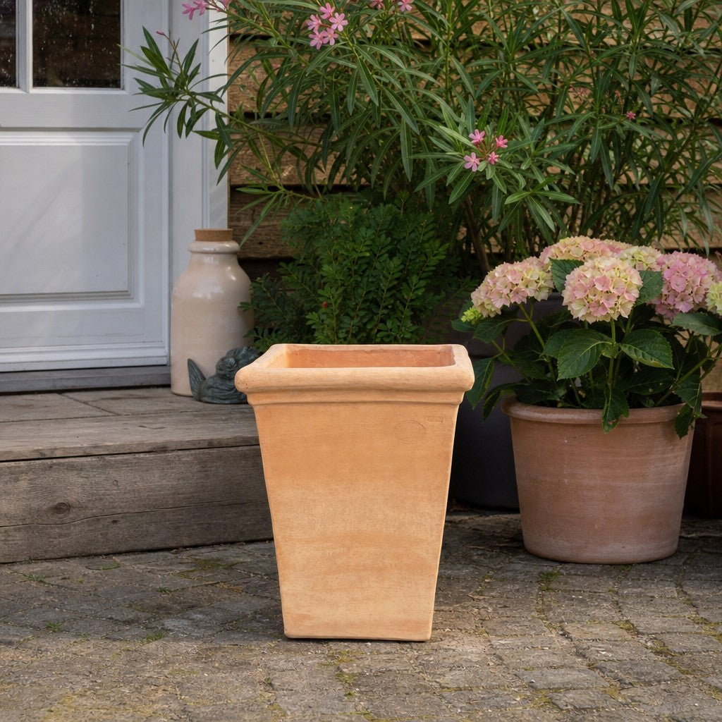 Auf einer Steinterrasse in der Nähe einer Holztreppe steht ein quadratischer Terrakotta-Blumenkübel, umgeben von Topfpflanzen mit rosa und weißen Blüten, im Hintergrund ist eine weiße Tür zu sehen.