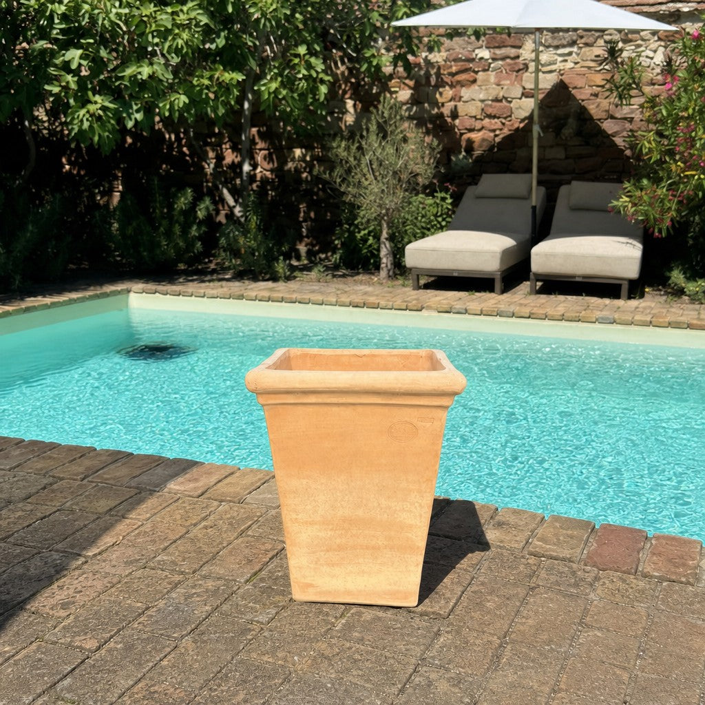 Auf einer gemauerten Terrasse neben einem Swimmingpool steht ein quadratischer Terrakotta-Pflanzkübel; im Hintergrund sind zwei Liegestühle und ein Sonnenschirm zu sehen, umgeben von Grünpflanzen und einer Steinmauer.