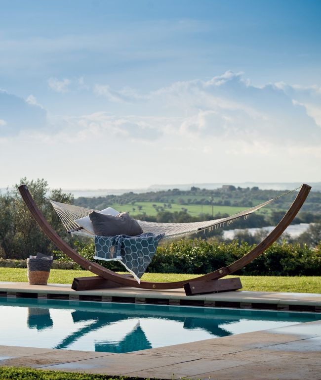 Eine Hängematte komplett mit Netzwerk, komplett mit Plüschkissen und einer Decke, hängt an einem Holzständer neben einem ruhigen Swimmingpool. Der Hintergrund zeigt eine üppige, grüne Landschaft und einen leicht bewölkten blauen Himmel, was eine friedliche Umgebung im Freien suggeriert – vollkommene Entspannung vom Feinsten.