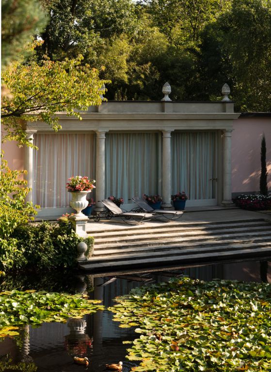 Eine ruhige Gartenszene mit einem luxuriösen Gebäude mit gewölbten Türen und Säulen. Die Terrasse, geschmückt mit dem grafitgrauen „Liege Aurora“ mit angenehmer Bespannung, ist mit Liegestühlen und Blumentöpfen geschmückt. Im Vordergrund befindet sich ein Teich voller Seerosen und schwimmender Enten, umgeben von Bäumen und Grünpflanzen. Die friedliche Umgebung ist in das sanfte Licht der Aurora getaucht.