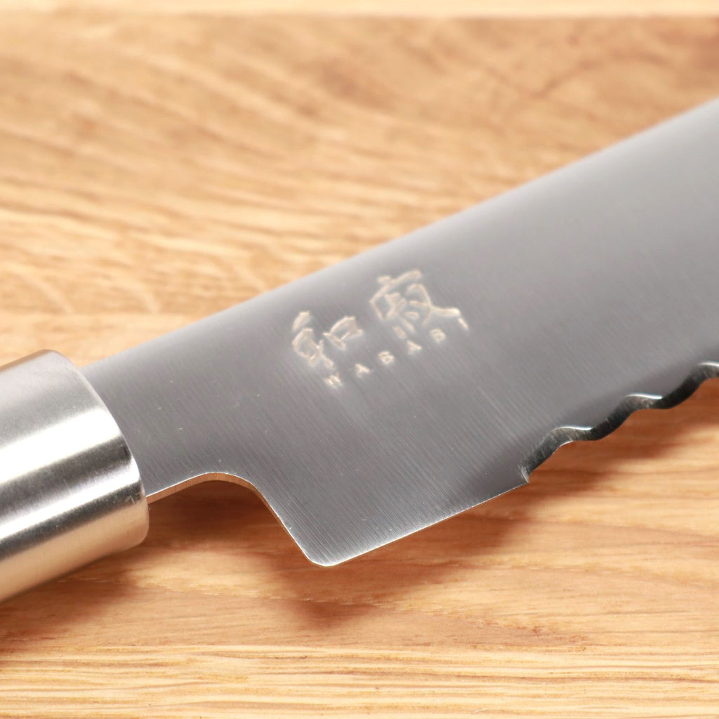 Nahaufnahme eines gezackten Kai Wasabi Black Brotmessers mit einer 23 cm langen Klinge auf einem großen Eichen-Schneidebrett 40x30 cm. Das Messer hat eine Klinge aus rostfreiem Stahl, in deren Nähe japanische Schriftzeichen eingraviert sind. Der Griff ist aus Metall und hat eine glatte Oberfläche.