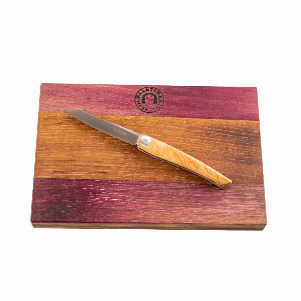 Ein farbenfrohes, rechteckiges, handgefertigtes Fassholzbrett mit auffälligen violetten, gelben und braunen Streifen verfügt über ein Nesmuk Soul Folder-Messer mit hellem Holzgriff und einer sorgfältig auf dem Brett platzierten Edelstahlklinge. Das Schneidebrett zeigt außerdem oben in der Mitte ein kreisförmiges Logo.