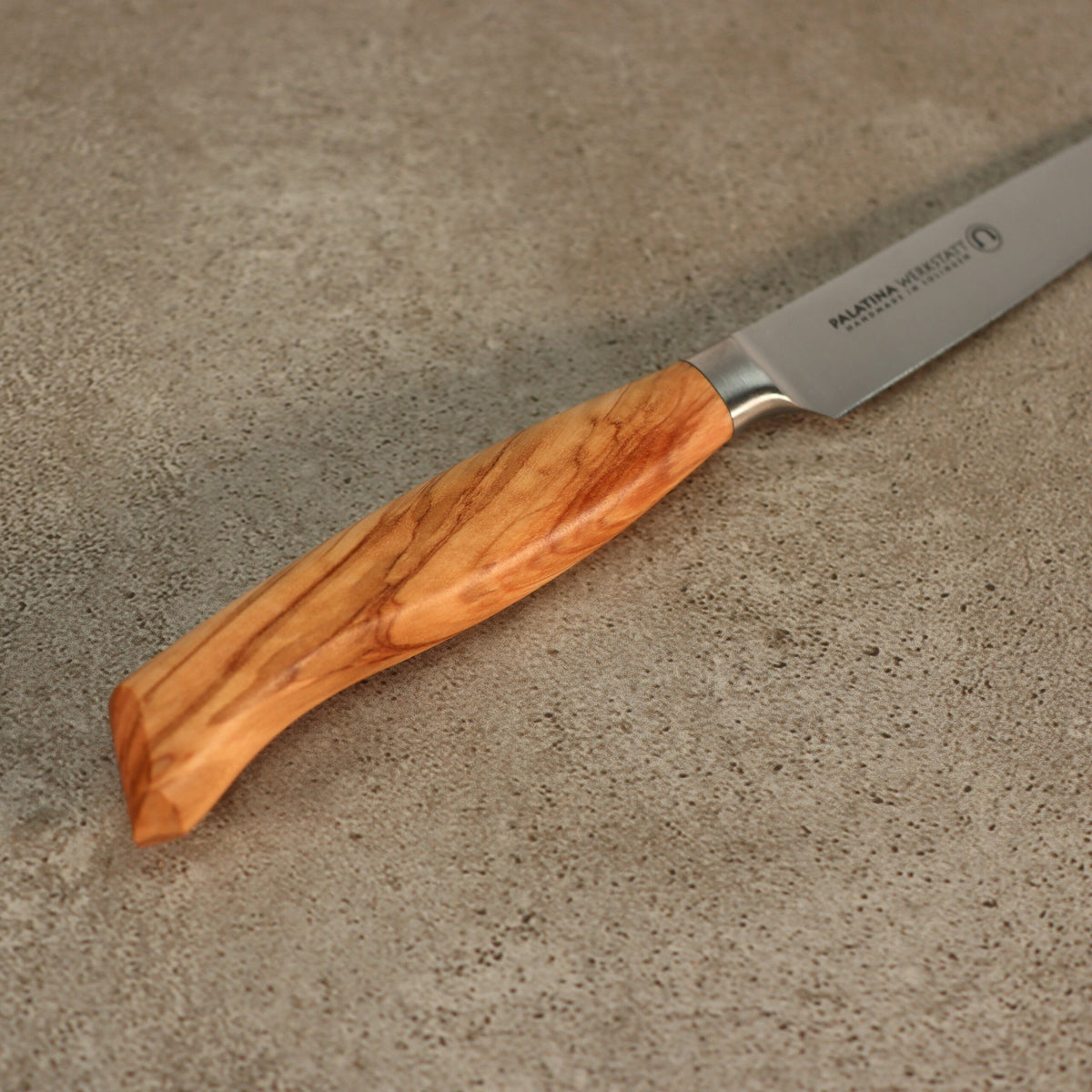 Ein Palatina Werkstatt Messerserie Officemesser 12 cm, Olivenholzgriff liegt auf einer Betonoberfläche. Das Messer hat eine glänzende Klinge aus rostfreiem Stahl und einen glatten, polierten Holzgriff mit natürlicher Maserung. Der Olivenholzgriff unterstreicht seine Eleganz, während der eingravierte Text auf der Klinge auf seinen Ursprung bei Palatina Werkstatt hinweist.